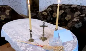 Dwa płonące świeczniki, krzyż, kropidło i białe kartki na stole przykrytym koronkowym obrusem.