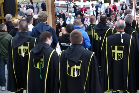 Grupa mężczyzn w czarnych płaszczach z żółtym symbolem krzyża i litery M.
