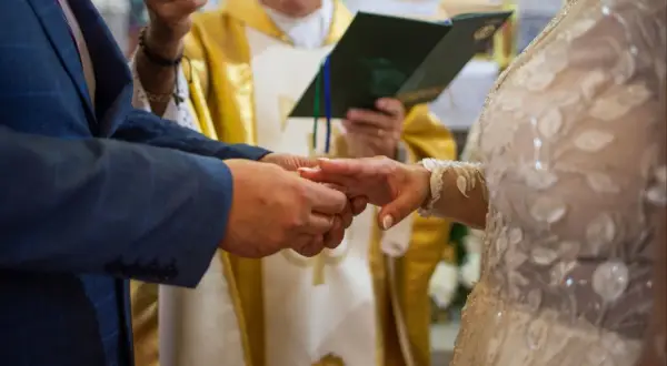Para młoda wymienia obrączki podczas ceremonii ślubnej, z księdzem w tle.
