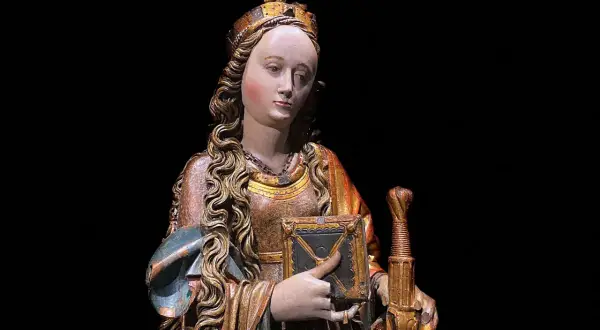 Drewniana figura świętej z koroną, księgą i mieczem na czarnym tle.