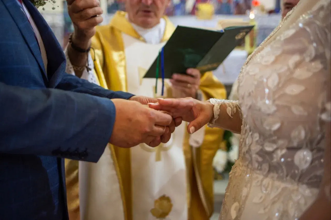 Para młoda wymienia obrączki podczas ceremonii ślubnej, z księdzem w tle.