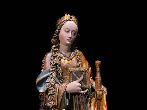 Drewniana figura świętej z koroną, księgą i mieczem na czarnym tle.