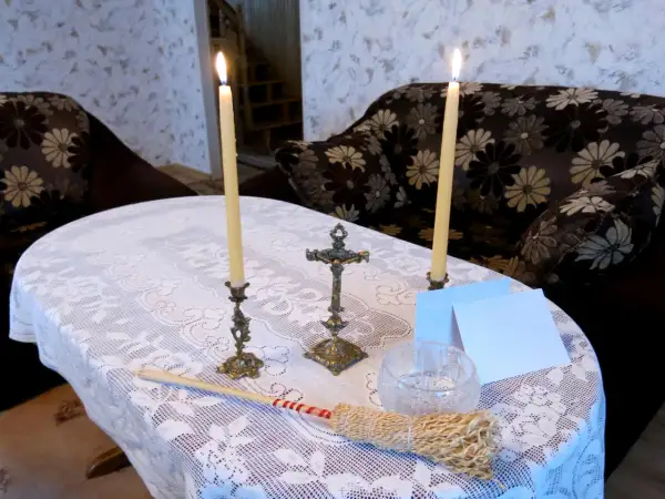 Dwa płonące świeczniki, krzyż, kropidło i białe kartki na stole przykrytym koronkowym obrusem.